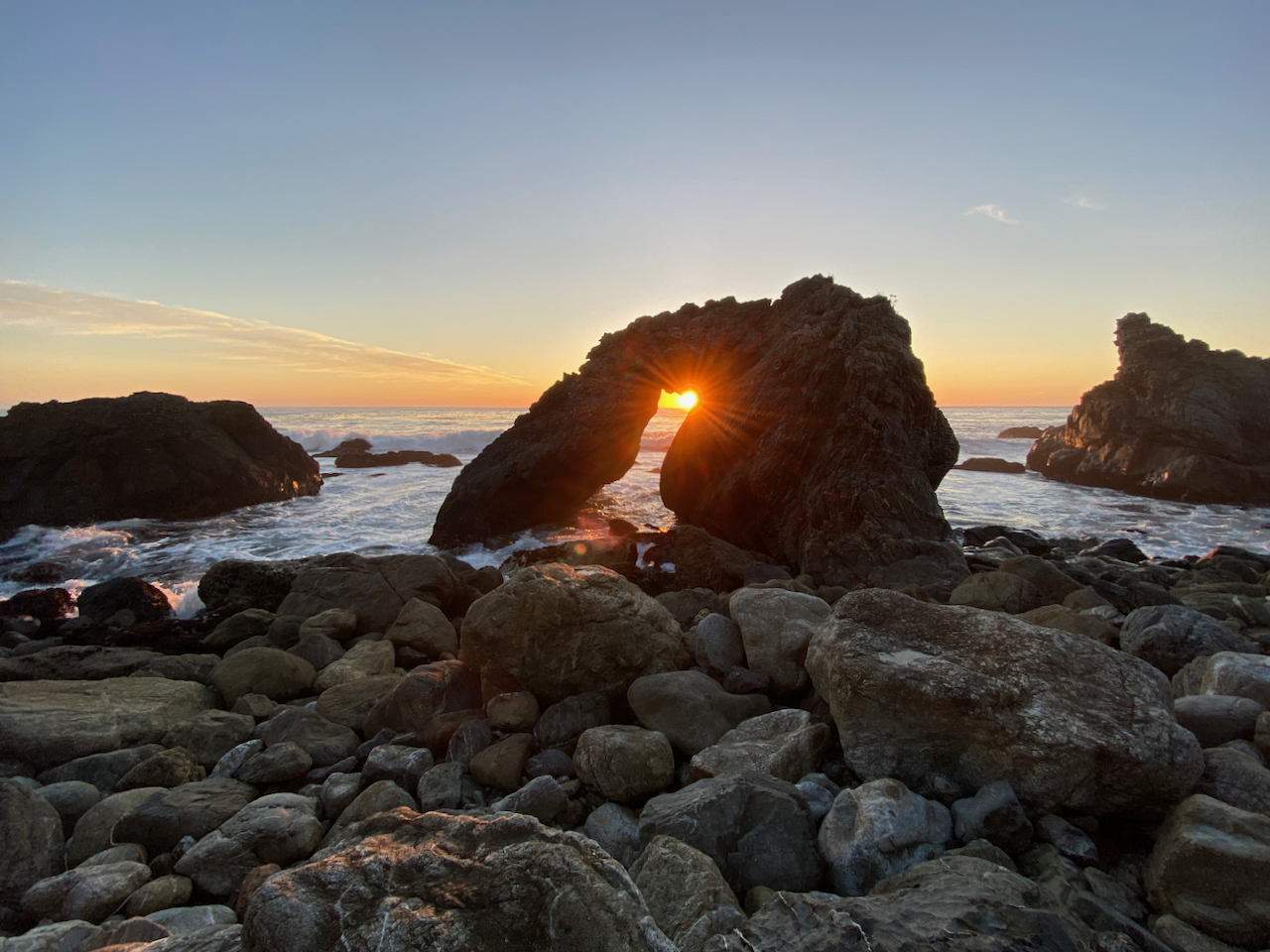 Sun setting through a rock arch with waves crashing in the distance.